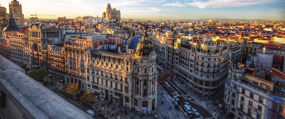 Vista aérea de la Gran Vía de Madrid, destacando la cúpula negra y dorada del Edificio Metrópolis en la intersección, con el horizonte de la ciudad extendiéndose bajo un cielo despejado.